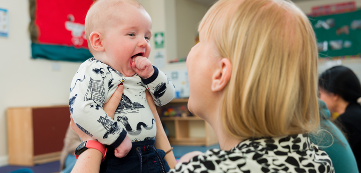 Mother holding her baby up to her face and smiling. The baby has their mouth wide open with their fist near their mouth.