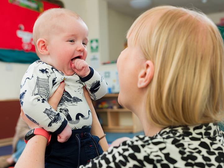 Mother holding her baby up to her face and smiling. The baby has their mouth wide open with their fist near their mouth.