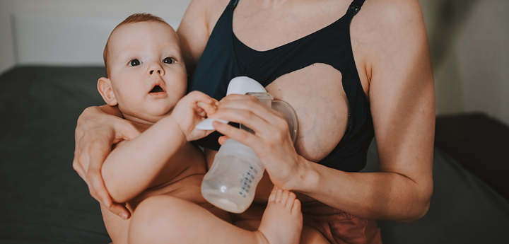 Woman using a pump in one hand to express milk whilst holding her baby in her other arm