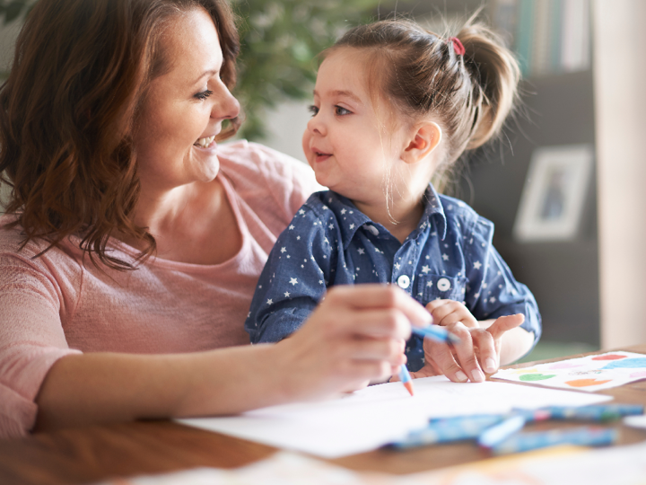 Young girl sitting on an older woman's lap, smiling at each other. They are both holding a crayon and a piece of paper is in front of them on a table