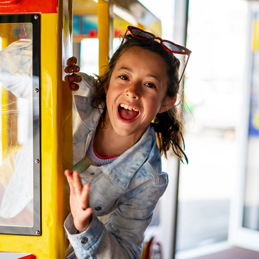 Young girl with sunglasses on her head leaning out of a supermarket ride whilst smiling with mouth wide open.
