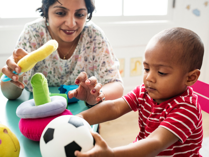 Toddler sitting at a child's table holding a football. A smiling adult is also sitting at the table and playing with the other soft toys on the table. 