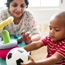 Toddler sitting at a child's table holding a football. A smiling adult is also sitting at the table and playing with the other soft toys on the table. 