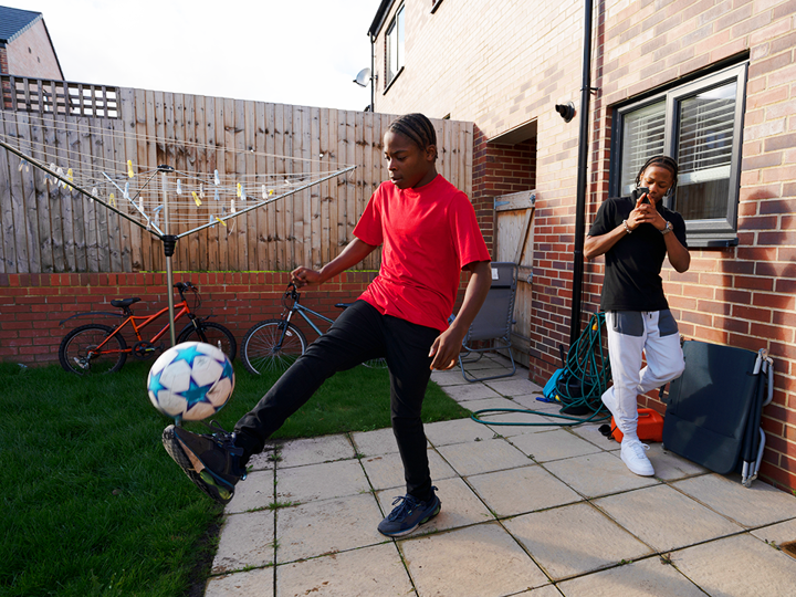 Teenage Boy Playing Kicking Ball In Back Garden on a stone patio with their male friend behind filming on their phone.