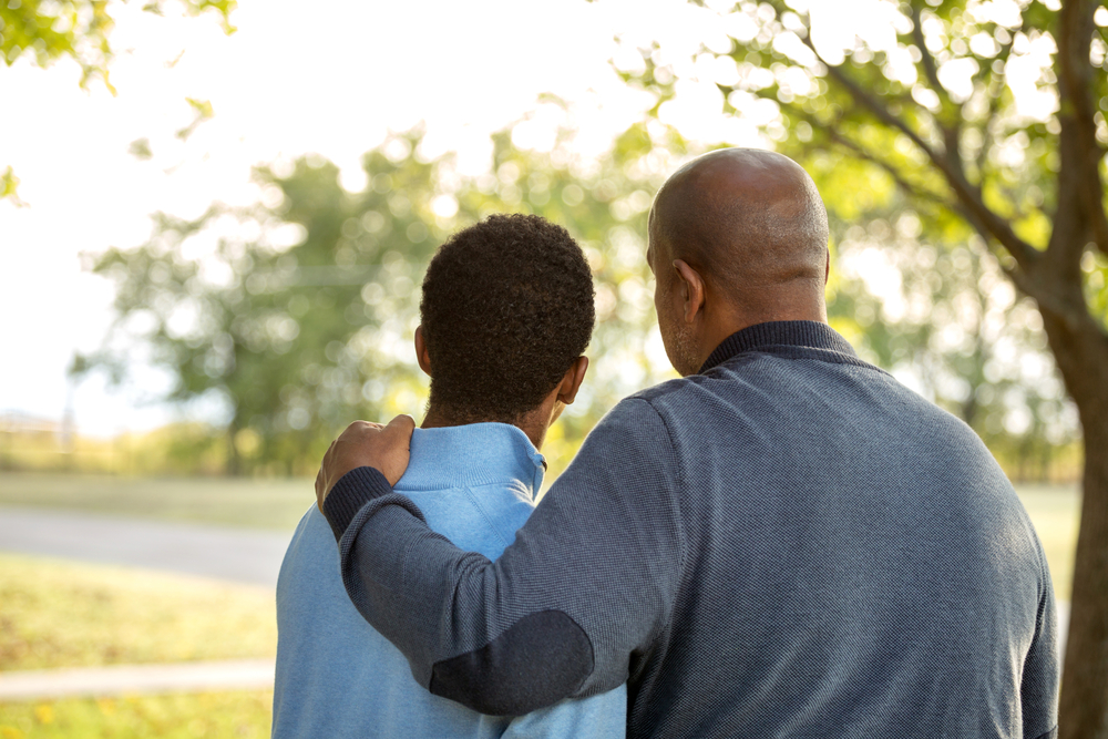 Teenage boy and dad standing with their back to the camera. The dad has his arm around his son.