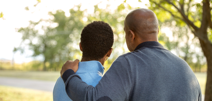 Teenage boy and dad standing with their back to the camera. The dad has his arm around his son.