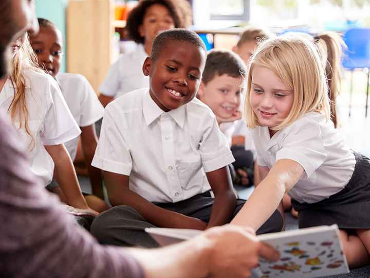 Children In A Class Looking At A Book Held By A Teacher