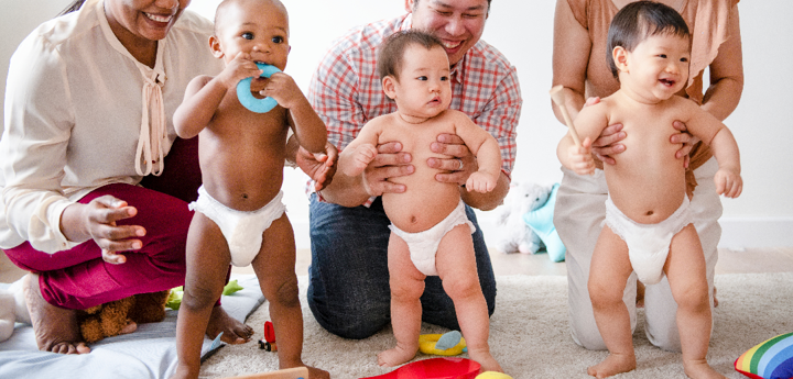 3 babies standing in a row in nappies, each being supported by an adult kneeling behind them