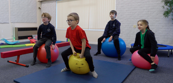 3 young boys and 1 young girl wearing school uniform and sitting on space hoppers in a classroom.