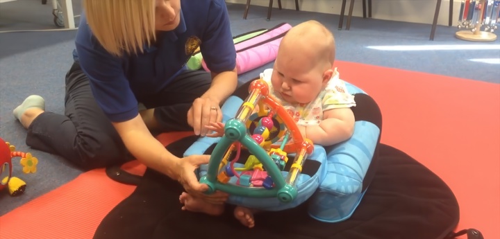 Baby Girl Sitting Up Using Support With An Adult Woman Placing A Toy In Their Lap