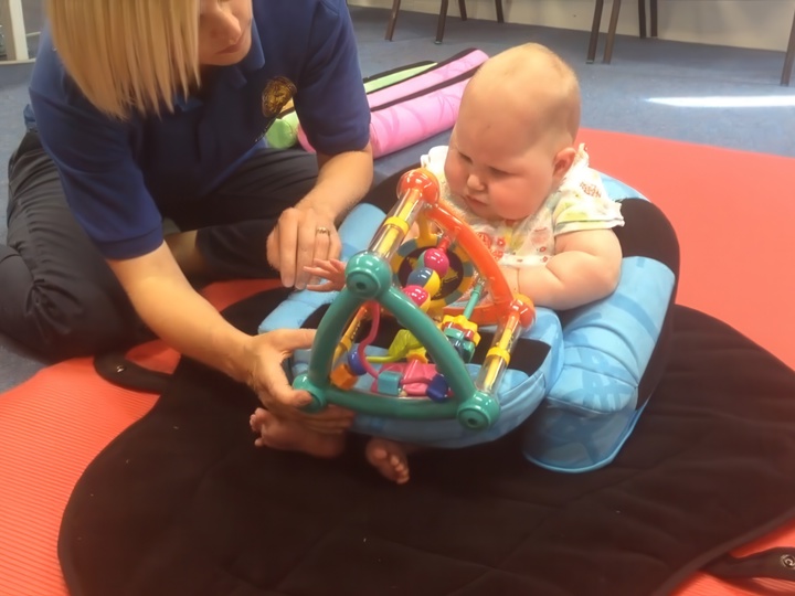 Baby Girl Sitting Up Using Support With An Adult Woman Placing A Toy In Their Lap