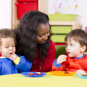 Two toddlers eating fruit sitting at a child's table in a nursery setting. A carer is sitting behind them. 