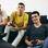 3 teenage boys, boy 1 (left) is laughing and holding a mug, boy 2(center) playing on playstation and concentrating, boy 3 (right) is playing on playstation and smiling