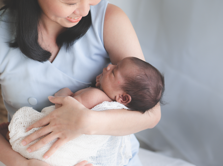 A woman sitting down and smiling whilst looking down at the baby she is holding, which is wrapped in a blanket, in her arms