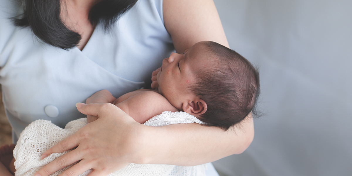 A woman sitting down and smiling whilst looking down at the baby she is holding, which is wrapped in a blanket, in her arms