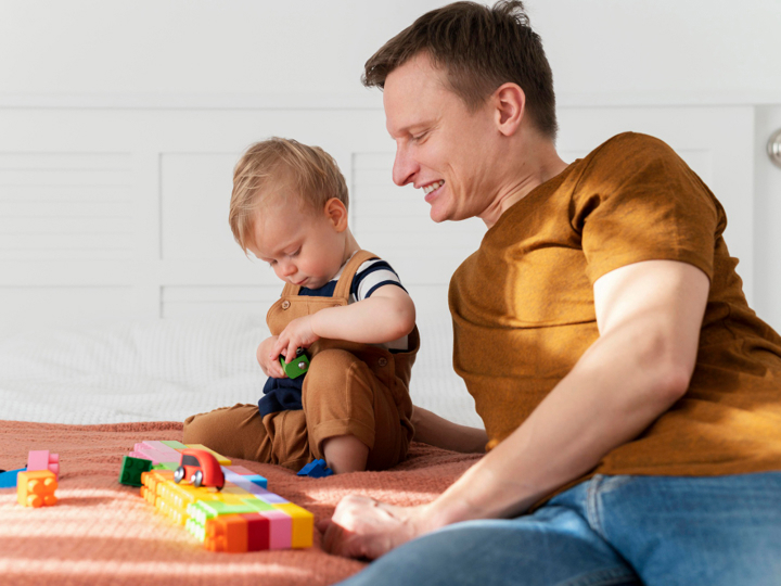 Father And Son Sitting On A Bed Playing With Coloured Blocks And Toy Cars
