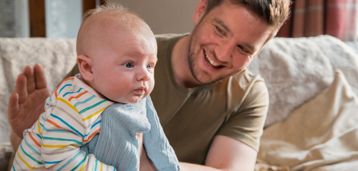 Dad sitting on the edge of sofa holding a baby and patting them on the back