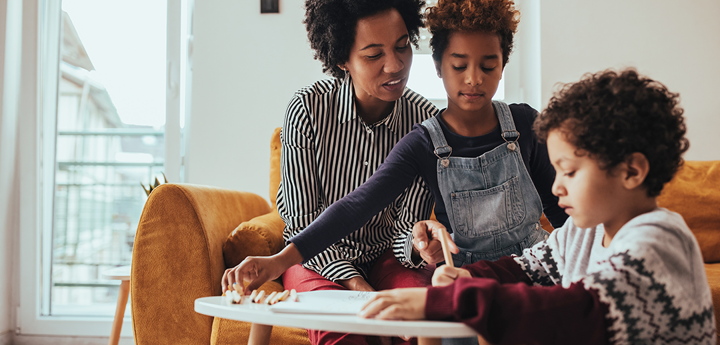 Adult woman and child sitting on a sofa looking at a young boy sitting on the floor writing on a piece of paper on a coffee table in front of them