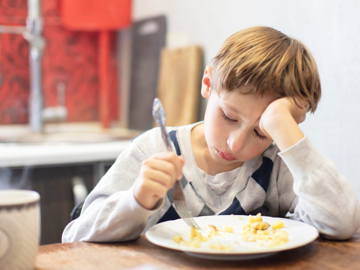 Young boy sitting at the kitchen table looking miserable. Holding a fork in one hand and his head is resting in his palm of the other hand.