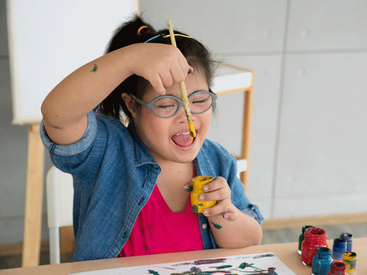 Young girl with glasses painting, sitting at a table dipping a paintbrush into a paint pot and smiling.