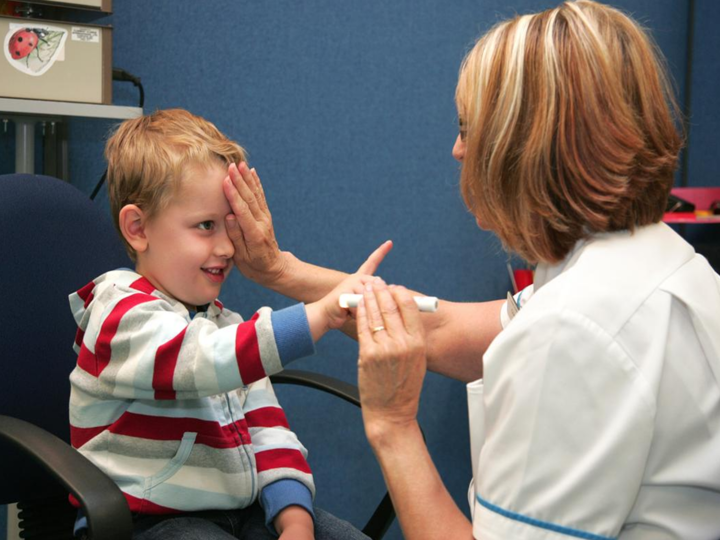 Young boy sitting in a chair with a clinician covering one of his eyes with the palm of her hand and holding a light in the other hand.