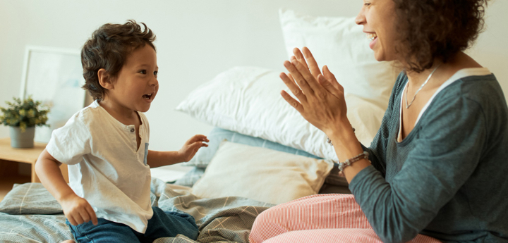 Mum and young son kneeling on the bed together. Both are smiling and look happy whilst mum is clapping her hands