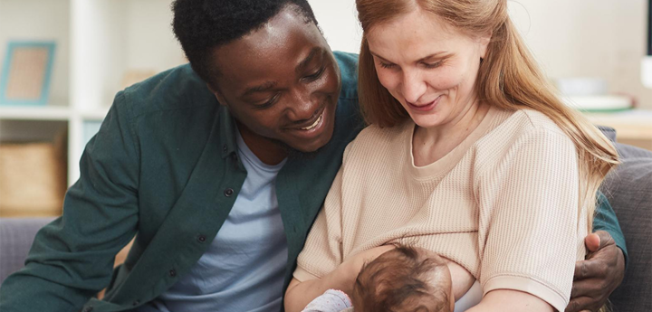 Mum sitting on sofa breastfeeding her baby with the dad sitting next to her with his arm round her. 