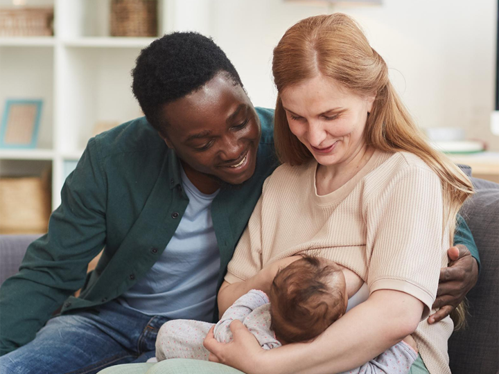 Mum sitting on sofa breastfeeding her baby with the dad sitting next to her with his arm round her. 