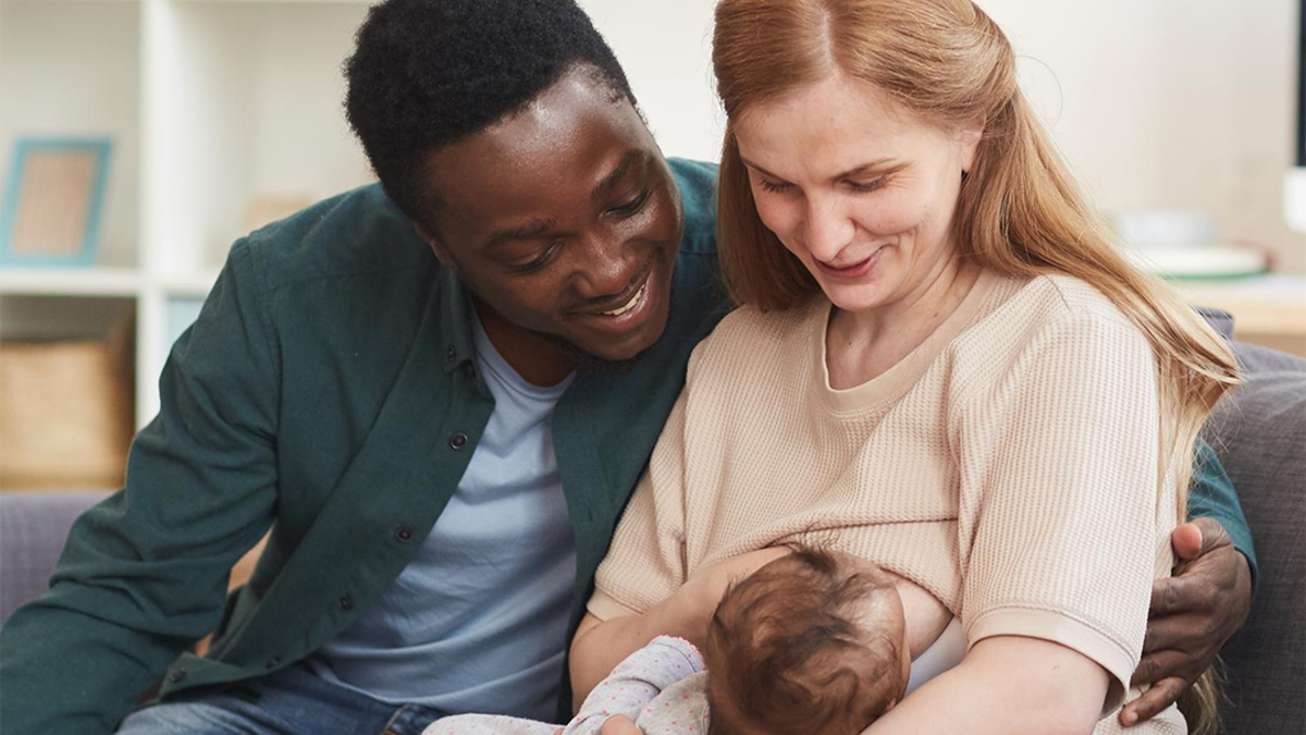 Mum sitting on sofa breastfeeding her baby with the dad sitting next to her with his arm round her. 