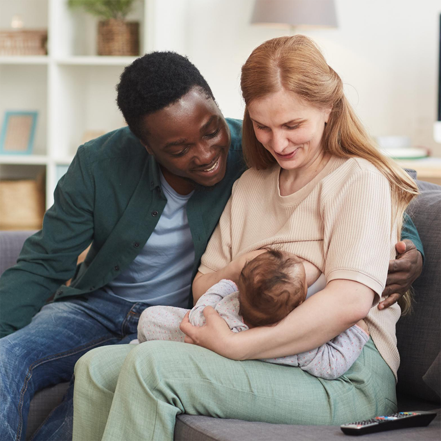 Mum sitting on sofa breastfeeding her baby with the dad sitting next to her with his arm round her. 