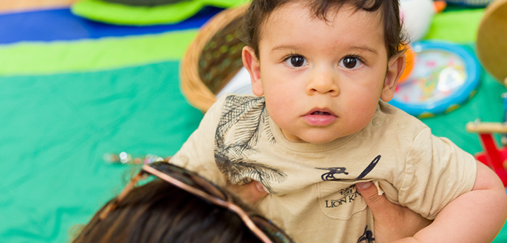 Toddler staring at the camera being held up by his mum who is sitting on the floor.