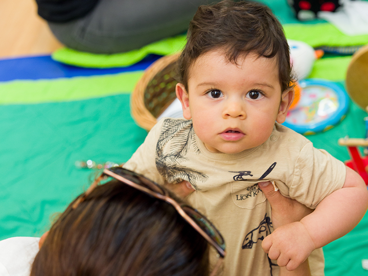 Toddler staring at the camera being held up by his mum who is sitting on the floor.