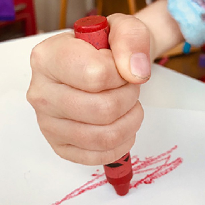 Child demonstrating the palmer grasp holding a red crayon and drawing on white paper