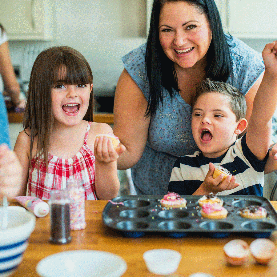 2 children and a woman making cupcakes at a kitchen bench. The boy is throwing his fist into the air excitedly whilst the girl is grinning.