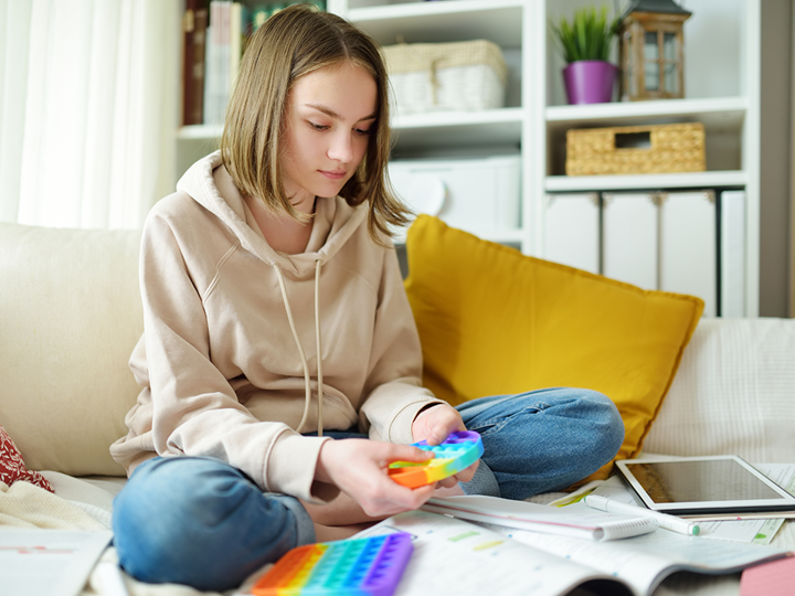 teenage girl on bed, legs crossed playing with fidget toy on bed with ipad and notebook in front of her