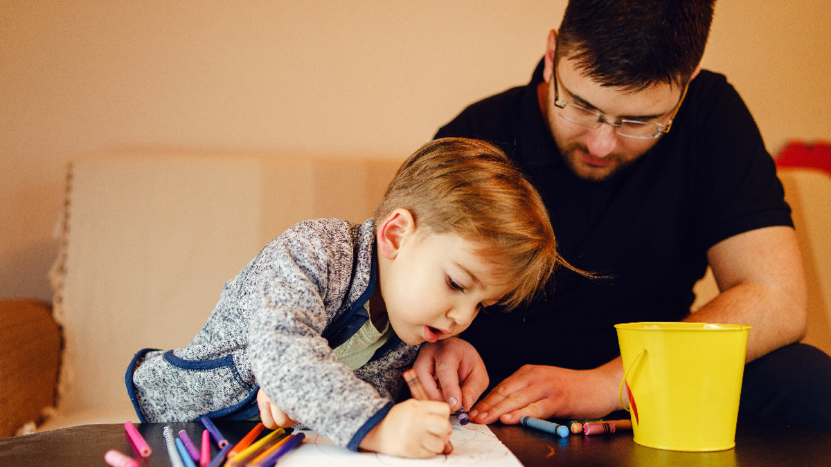 A child drawing with crayons and pencils on a coffee table whilst sitting on a sofa. Next to the child is their dad who is also drawing on the paper