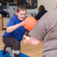 Young girl kneeling on an exercise mat balancing whilst holding an orange ball. An adult is kneeling in front for support.