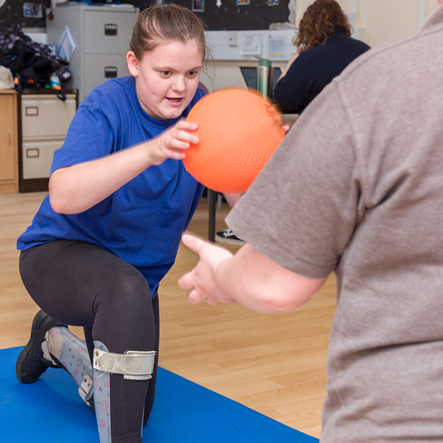 Young girl kneeling on an exercise mat balancing whilst holding an orange ball. An adult is kneeling in front for support.