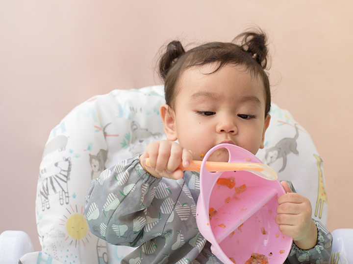 Baby in high chair playing with pink bowl with orange spoon