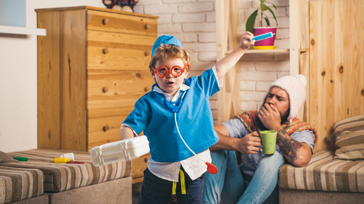 Child in nurses outfit with a toy stethoscope, toy syringe and a carrier box. Adult man in the background coughing and sat on the ground with a woolly hat and green mug