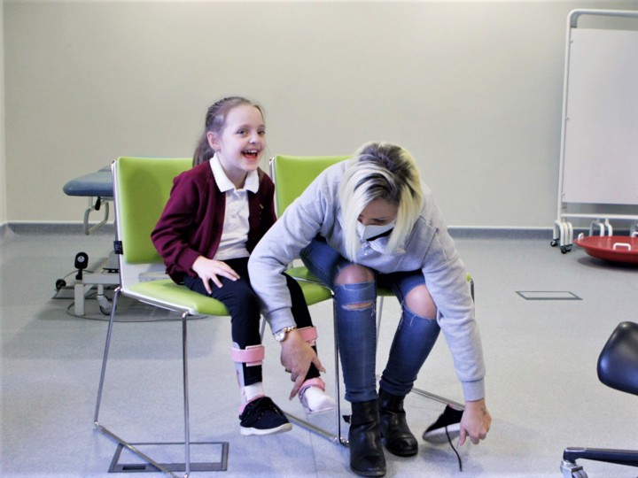 Young girl in a physio clinic sitting on a chair with an adult woman sitting next to her holding the girls leg and taking her shoe off.