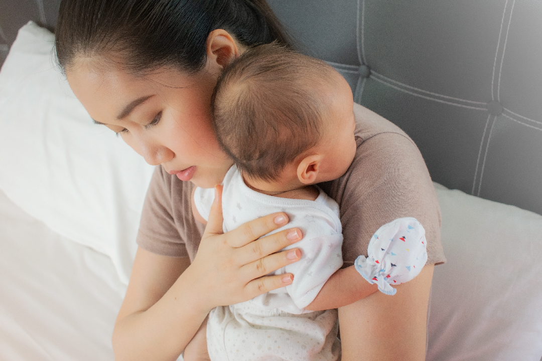 woman on bed holding baby with hand mitts against shoulder