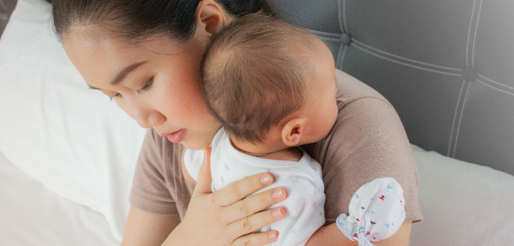 woman on bed holding baby with hand mitts against shoulder