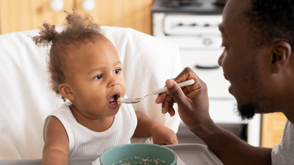 Dad sitting at a kitchen table with his baby girl in a highchair, feeding her food on a spoon