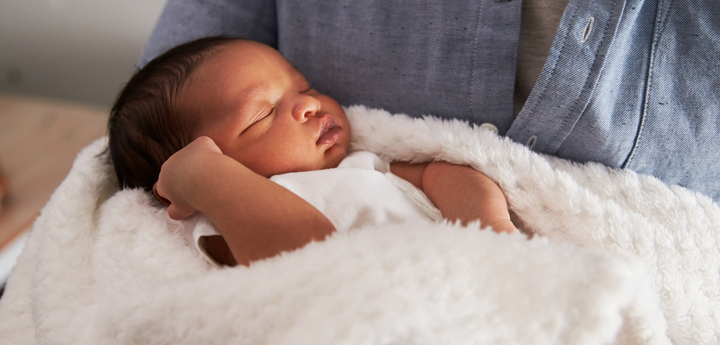 Baby asleep in a white blanket in the arms of an adult