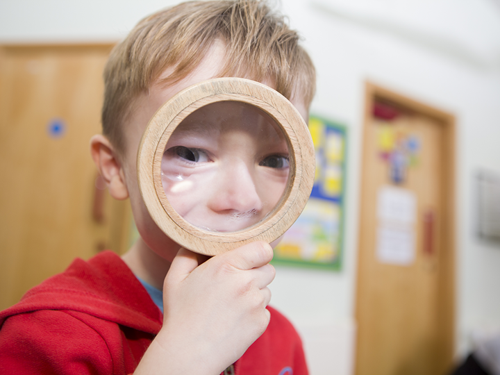 blonde haired young boy looking through wooden magnifying glass, his eyes and nose are exaggerated. background is a blurred cream wall with 2 wooden doors