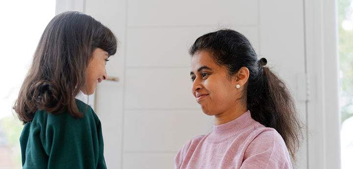 Adult kneeling in front of girl dressed in school uniform. Adult lightly touching child's arms, both smiling.