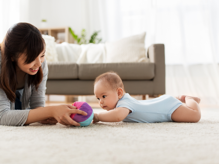 Mother and baby on floor with soft fabric ball. Baby is laying on their stomach on a rug, in a living room.