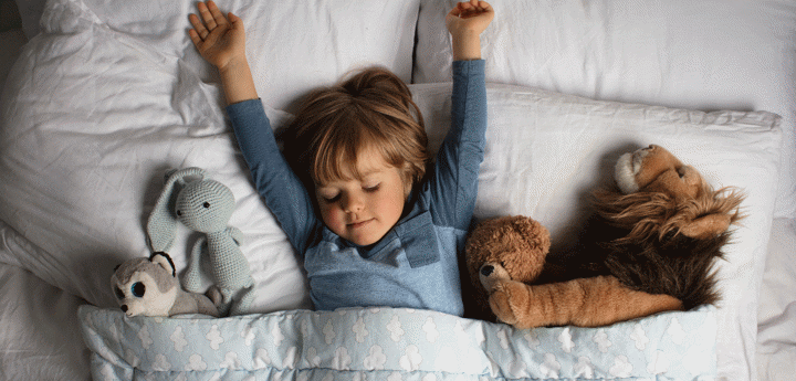 child lying in bed with their arms outstretched above them on the pillow and stuffed animals under the duvet either side of them