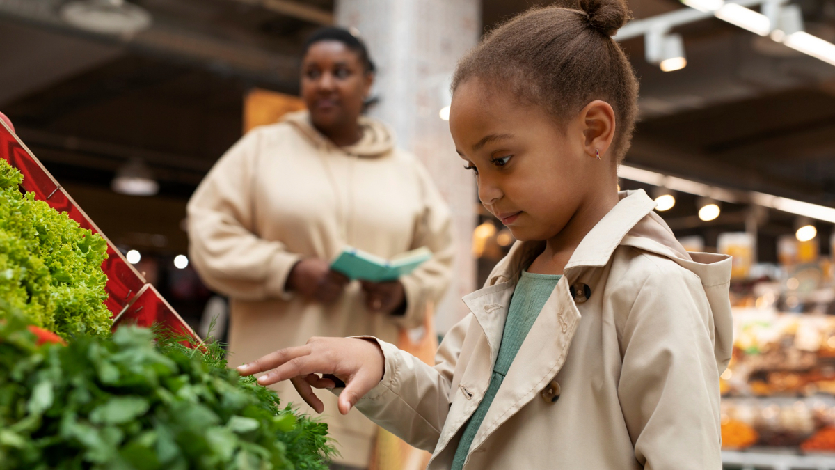 Young girl in a supermarket with hand outstretched looking at vegetables. In the background is her mother holding a shopping list